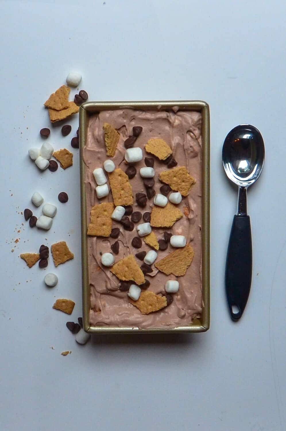 aerial picture of homemade s'mores ice cream in gold loaf pan with ice cream scoop and scattered graham crackers, chocolate, and marshamallows
