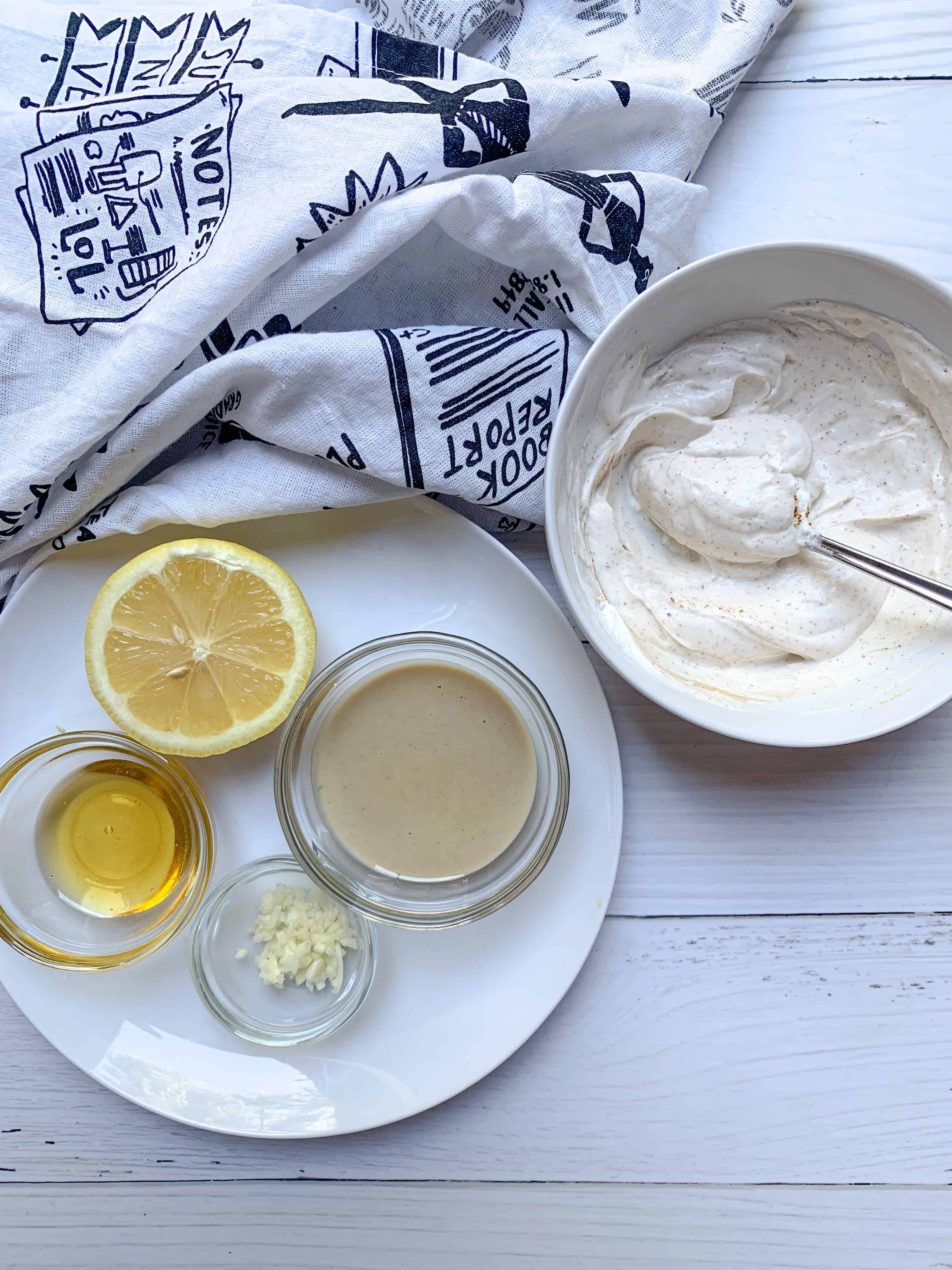 bowl of yogurt tahini dressing and plate with the ingredients to make next to it: with a lemon, honey, tahini, and garlic. 