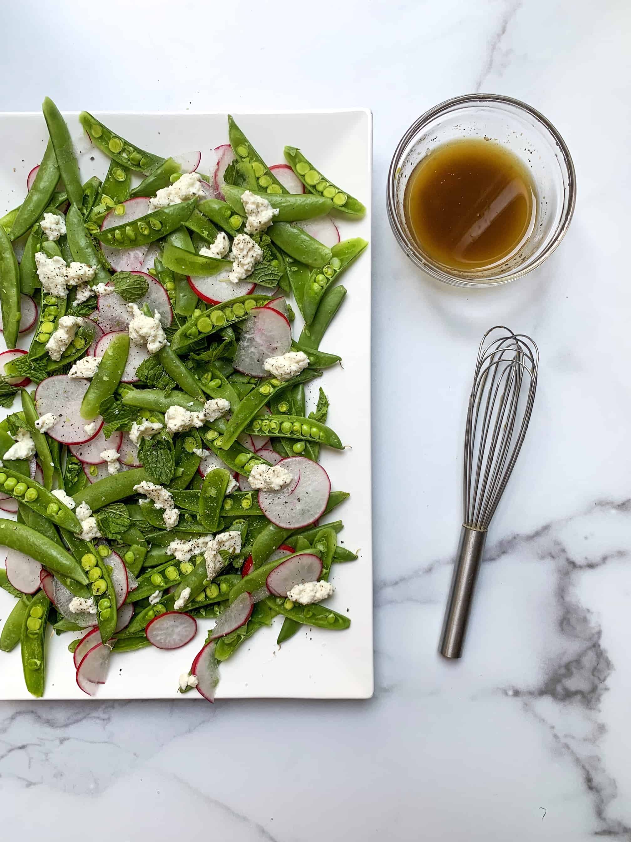 sugar snap pea salad on a white tray with bowl of lemon balsamic vinaigrette and whisk next to it