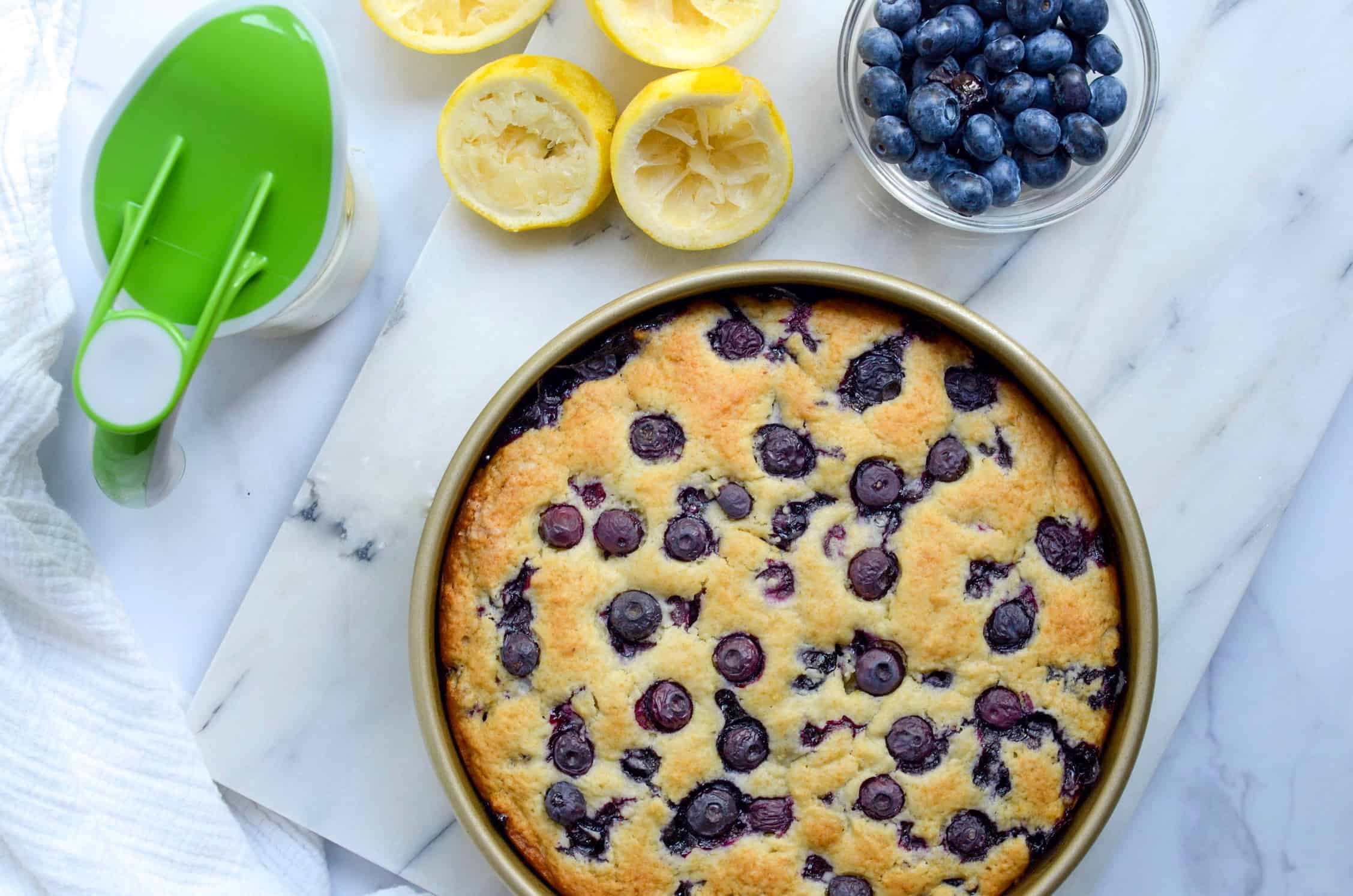 blueberry breakfast cake with lemon drizzle and fresh squeezed lemons and a bowl of blueberries. 