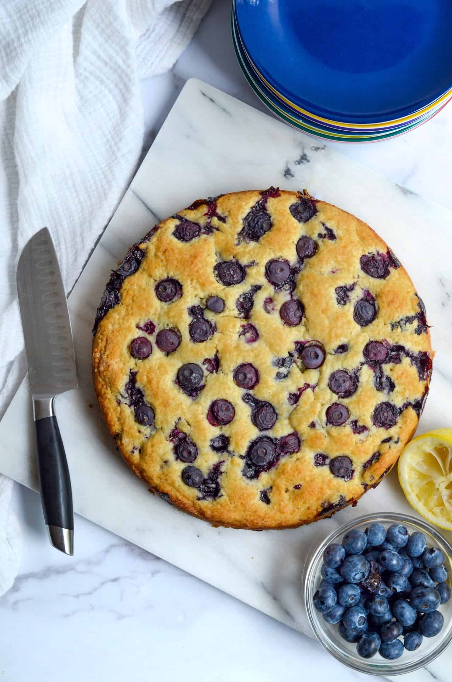 Aerial shot of freshly baked, round blueberry breakfast cake with a knife next to cake and a stack of plates.