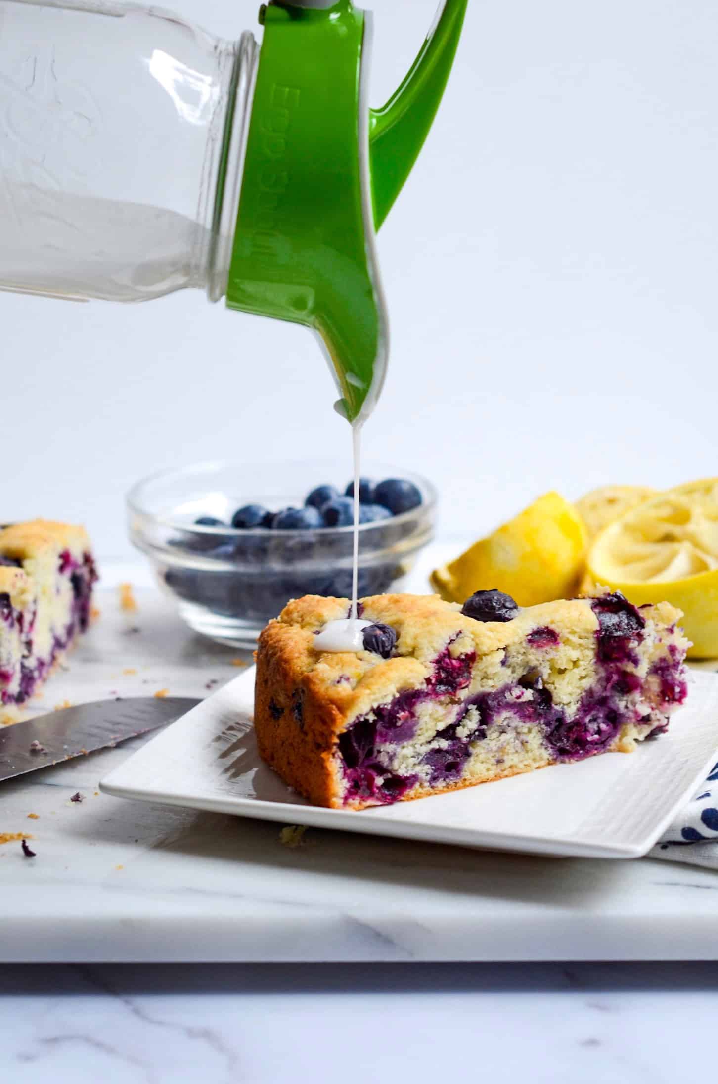 Pouring lemon drizzle over a slice of blueberry cake on a square white plate.