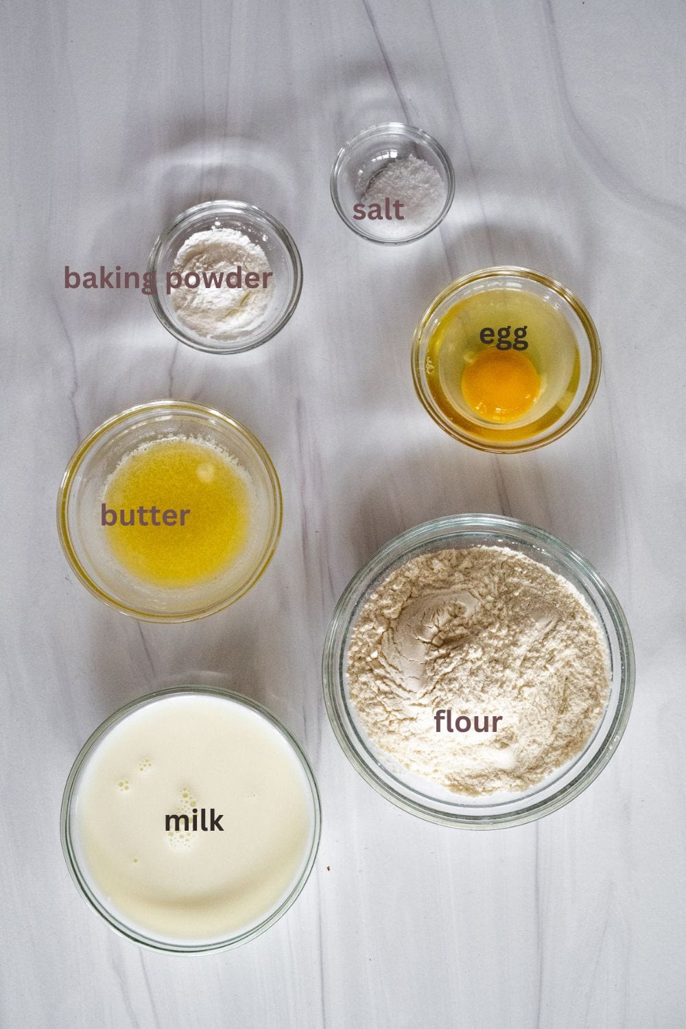 Individual ingredients for easy pancakes in individual bowls, including egg, baking powder, butter, flour, salt, and milk.