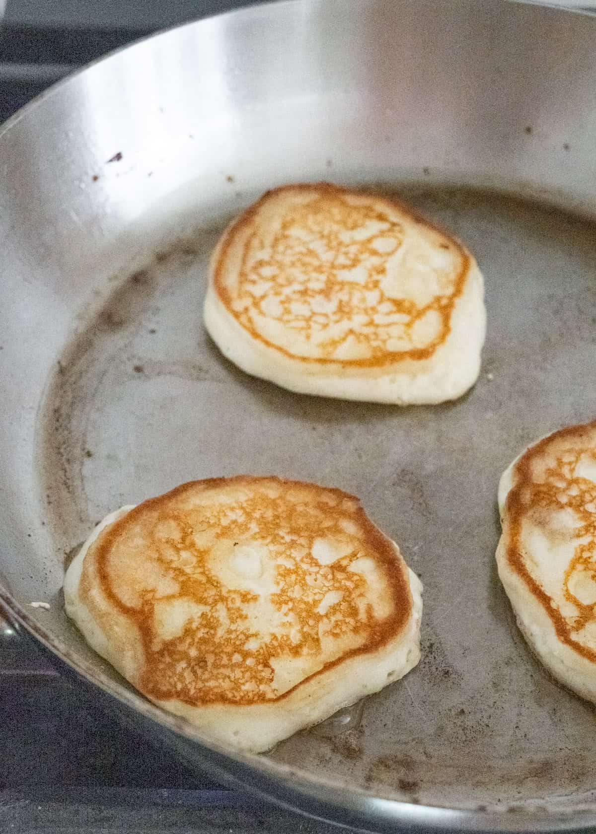 Three pancakes cooking in a stainless steel pan showing their golden tops.