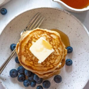 Aerial view of a stack of pancakes with a generous slab of butter with maple syrup and blueberries around the pancakes.