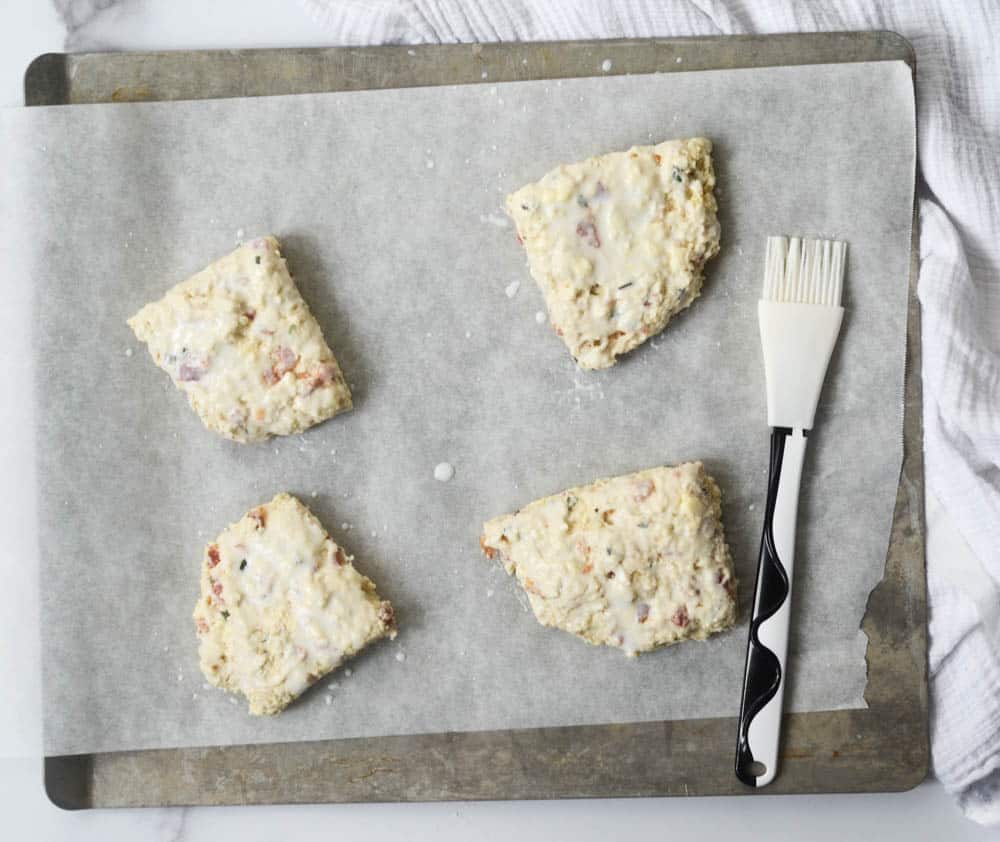 4 biscuits coated in buttermilk on baking sheet with pastry brush.