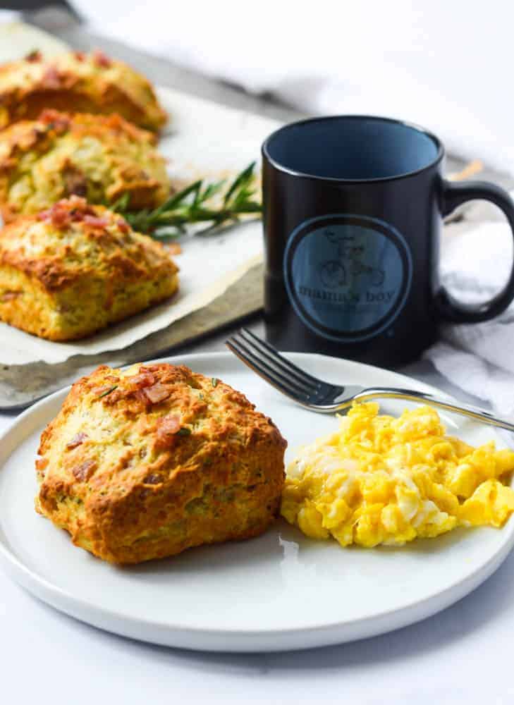 white plate with biscuit and scrambled eggs and fork.  mug and remaining biscuits in background on.baking sheet