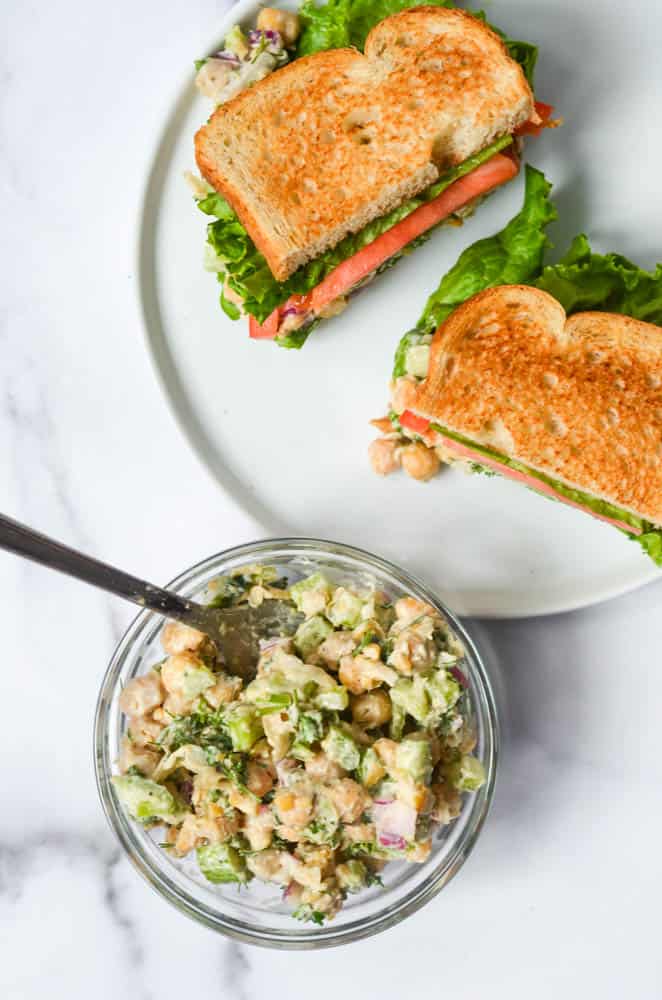 Overhead view of a glass bowl full of Easy Chickpea Salad with a sandwich in the background on a white plate