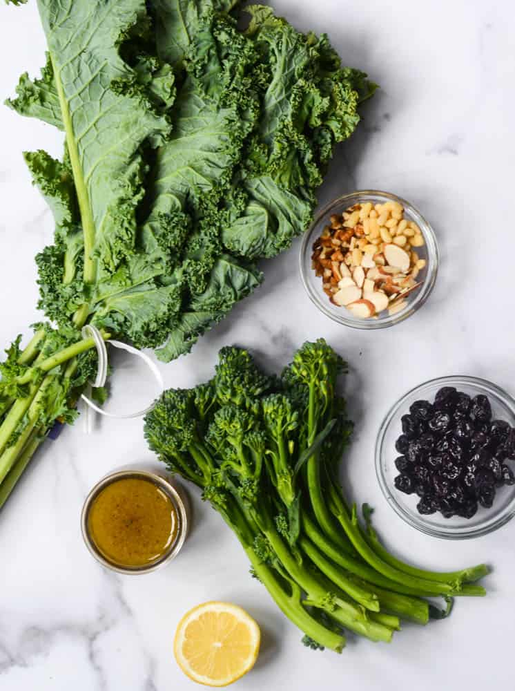 flat lay of all ingredients needed to make kale salad: kale, broccolini, nuts, sour cherries, lemon, and maple vinaigrette.