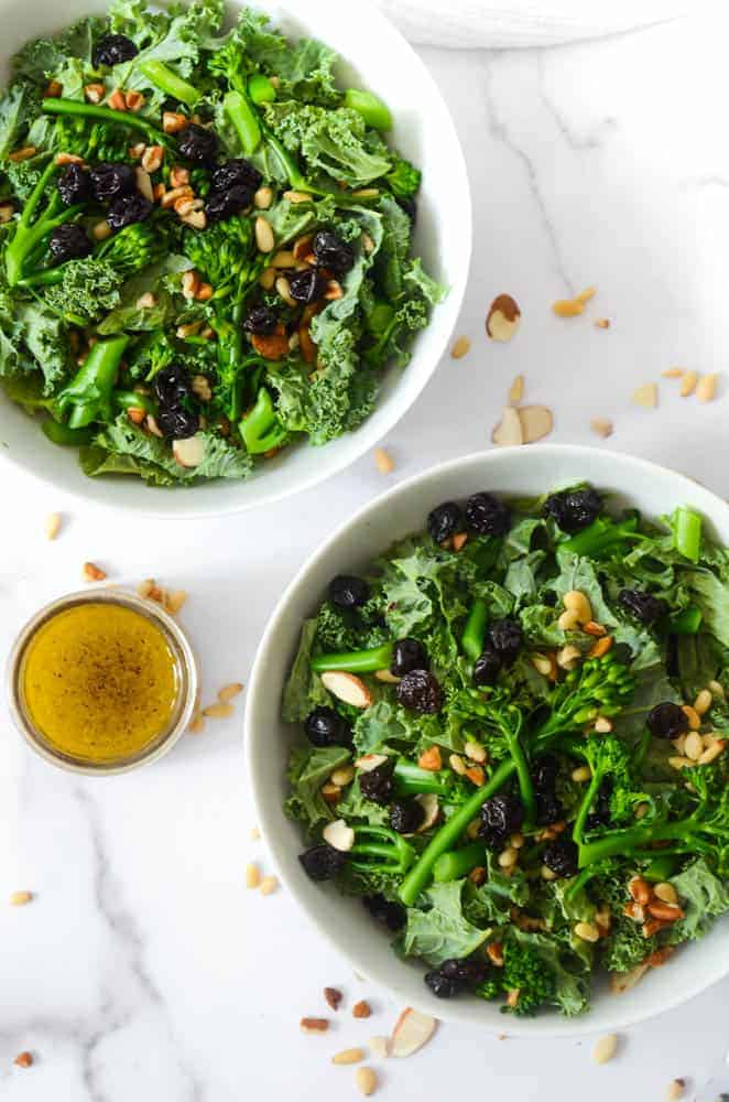 aerial shot of two bowls of kale salad with jar of vinaigrette and nuts scattered on the counter