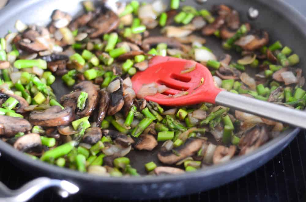 pan full of mushrooms, asparagus and onions with red spatula 