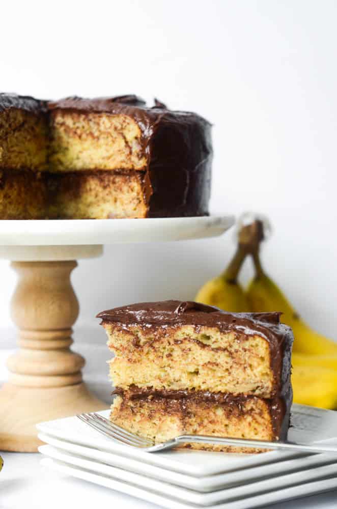 slice of banana cake on stack of white plates with rest of cake and bananas in the background
