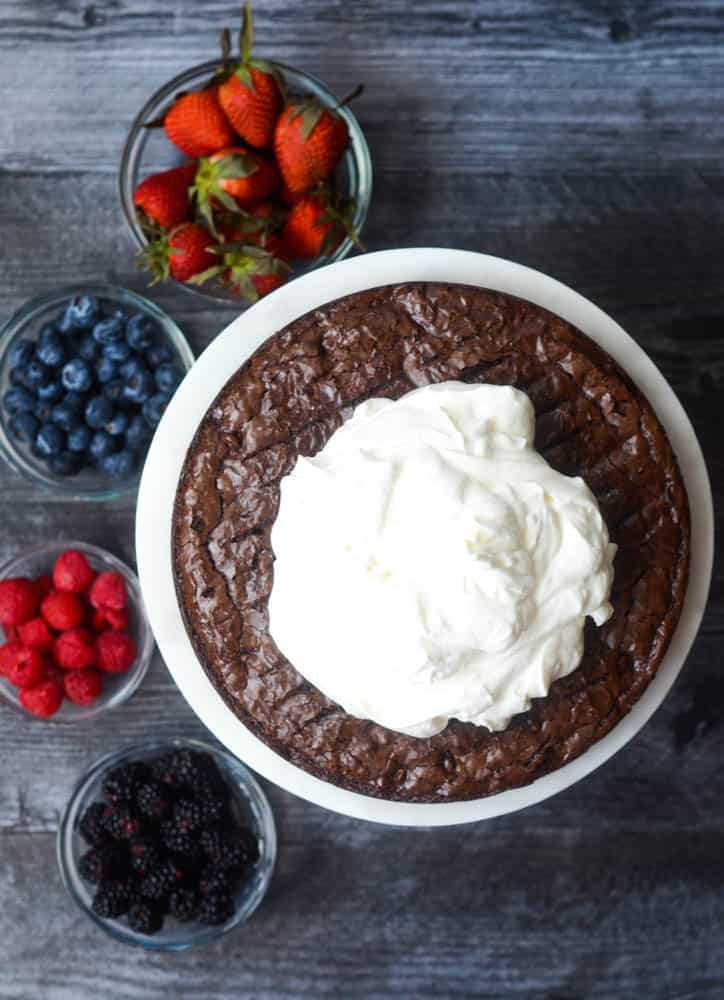 aerial view of brownie on cake plate with pile of whipped cream in the middle and bowls with different berries in them on black background