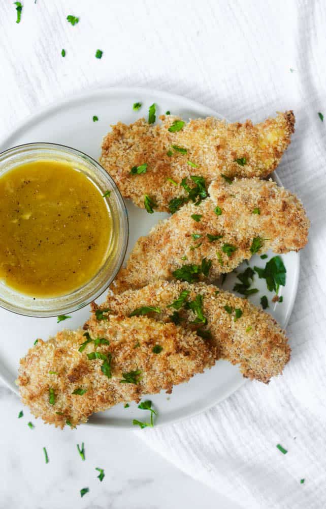 aerial view of baked buttermilk chicken tenders sprinkled with parsley with bowl of honey mustard