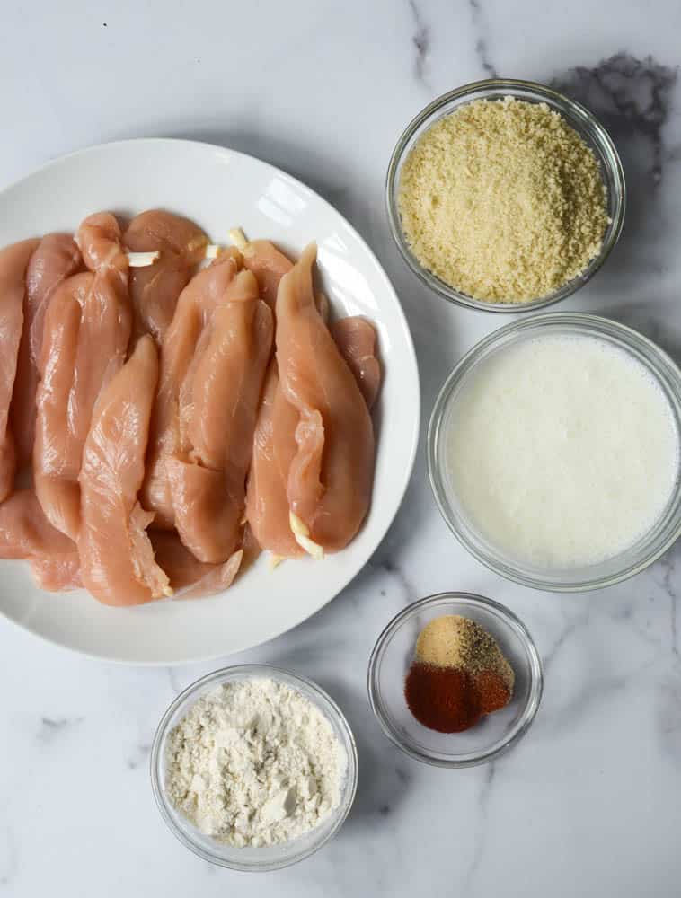 white plate with raw chicken on it, and bowls of breadcrumbs, buttermilk, spices, and flour. 