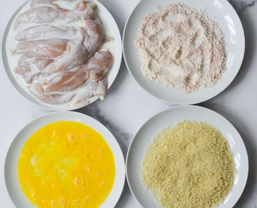 four white plates arranged in square with chicken tenders, flour mixture, eggs, and breadcrumbs. 