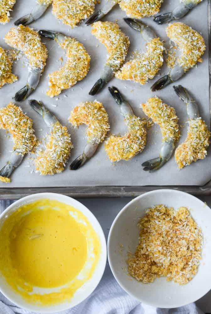 aerial view of tray of breaded shrimp with bowl of egg mixture and bowl of breadcrumbs and coconut. 