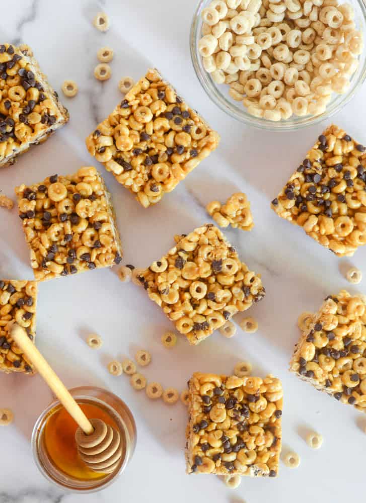 cut up cereal bar squares with jar of honey and bowl of cheerios on white background