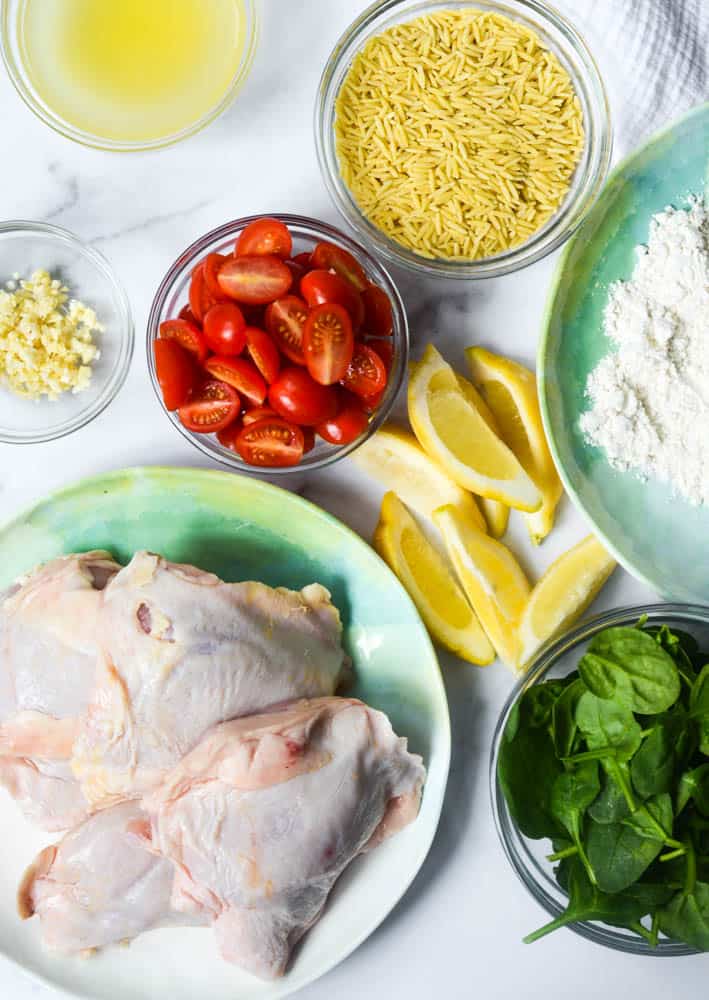 aerial shot of all ingredients needed: tomatoes, garlic, orzo, lemon, flour, spinach, and chicken thighs.
