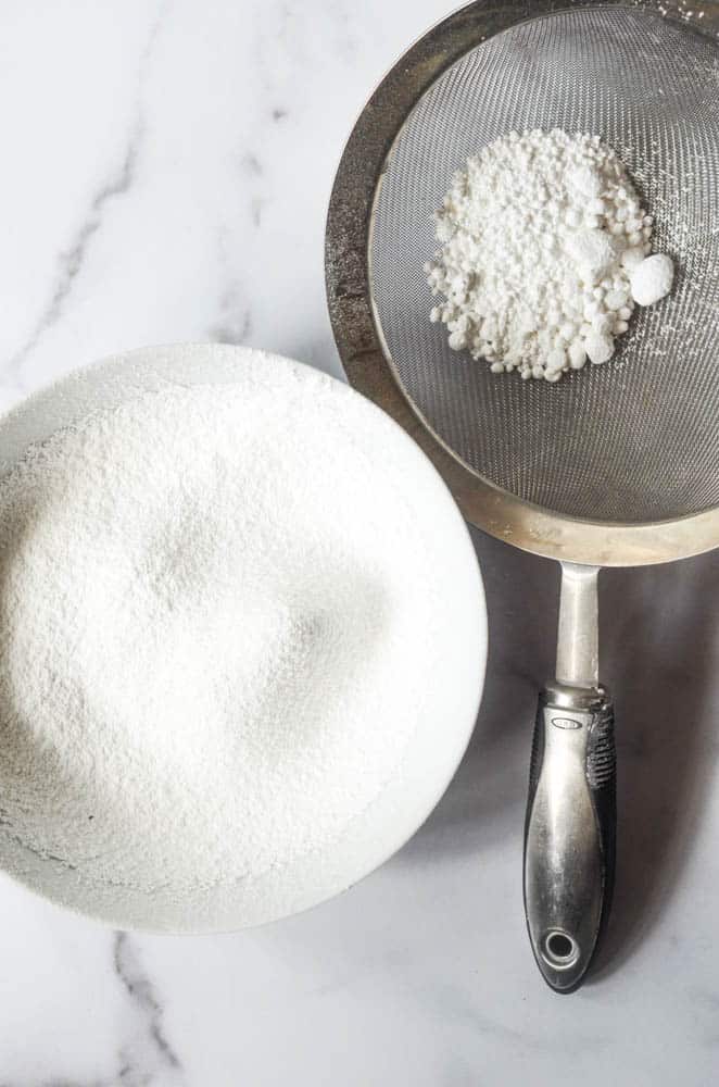 bowl of sifted confectioner's sugar next to sieve with unsifted confectioner's sugar