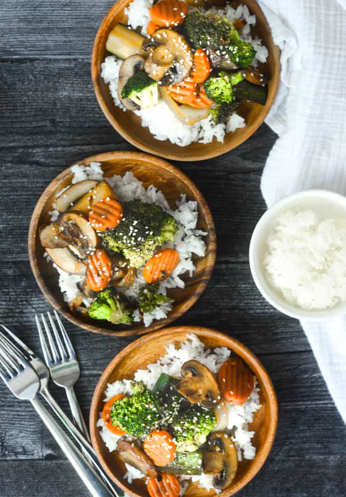 three wooden bowls of vegetables with forks and bowl of rice around bowls