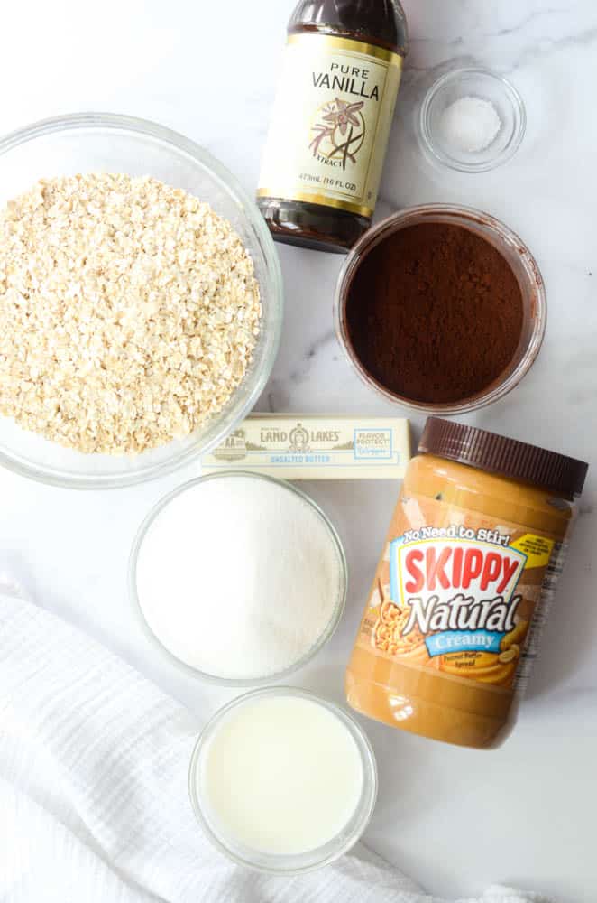 flat lay picture of ingredients needed to make no bake chocolate oatmeal cookies  on granite background - peanut butter, sugar, butter, milk, cocoa powder, vanilla extract and oats.