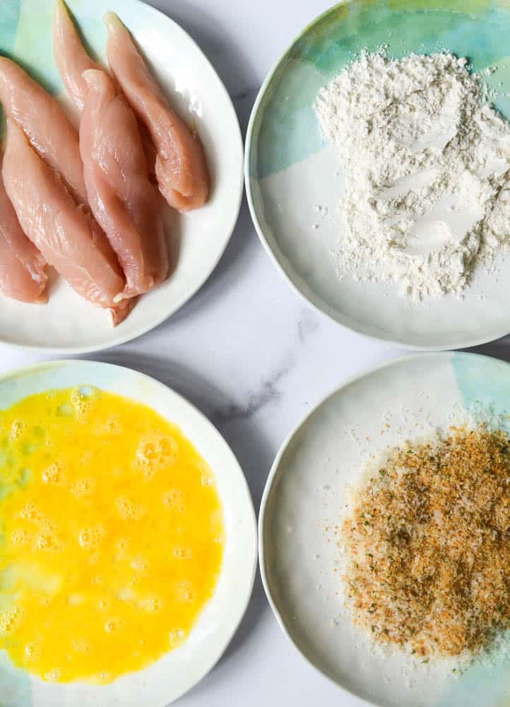 aerial view of four plates, one with chicken tenders, one with flour, one with eggs, and one with parmesan and breadcrumbs