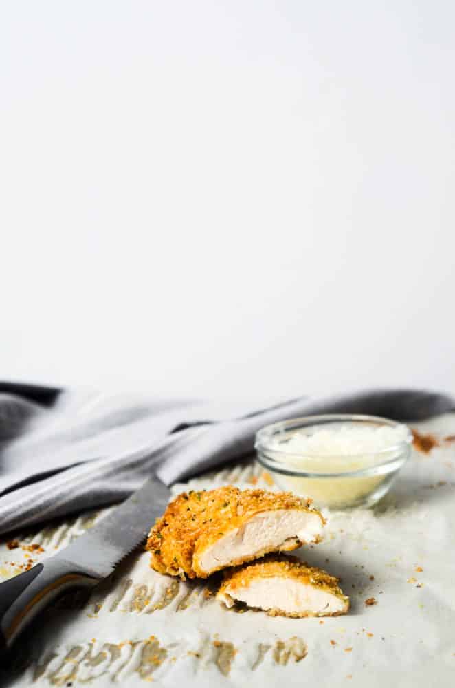 side shot of a parmesan chicken tender cut in half with inside exposed and a bowl of parmesan and knife in the background