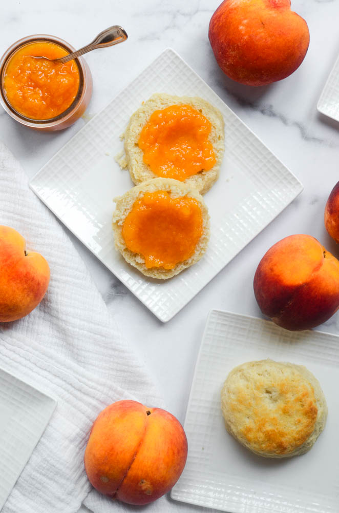 biscuit covered in peach jam with jar of peach jam to the side and peaches scattered on white background
