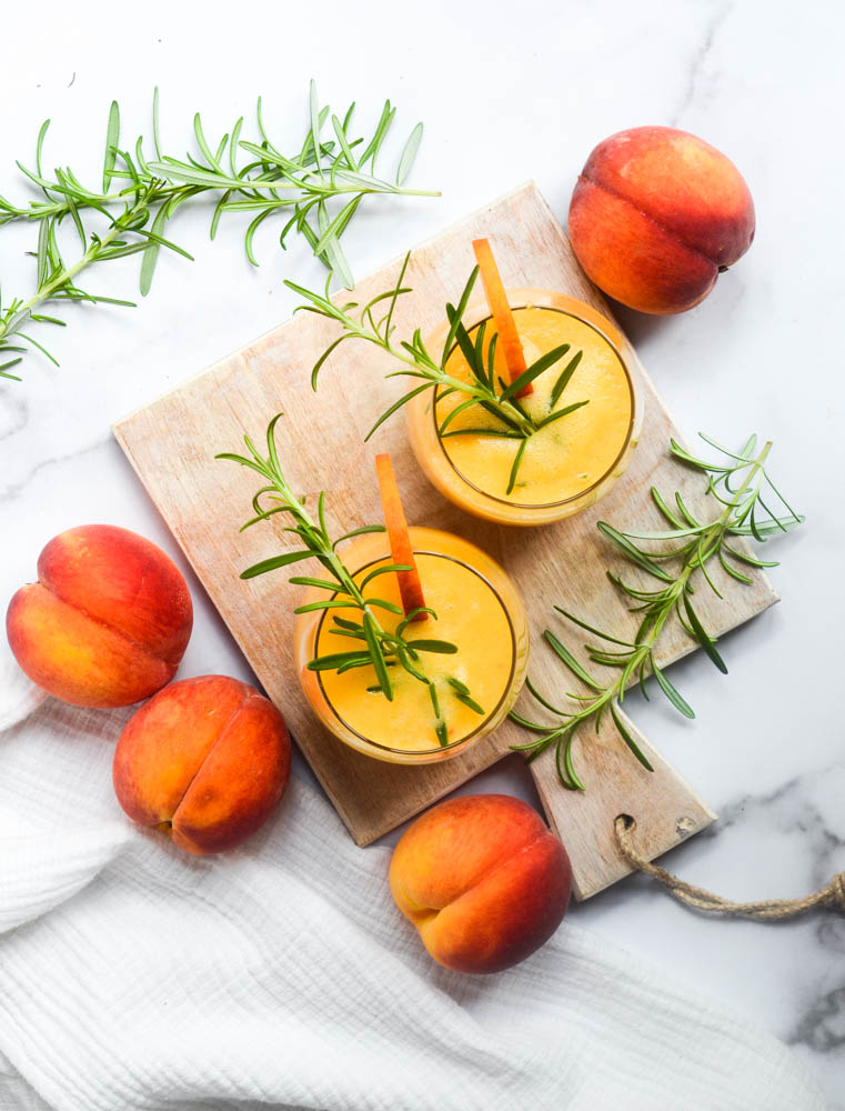 aerial picture of peach bourbon drinks on cutting board with sprigs of rosemary and peaches scattered around. 