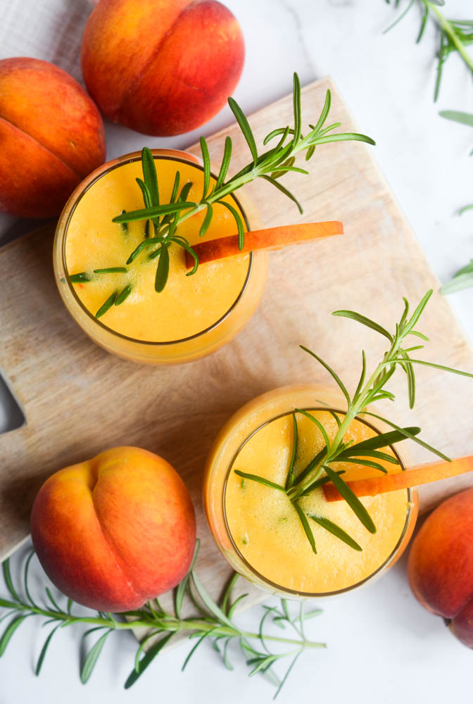 aerial picture of two peach bourbon cocktails on a wooden cutting board with a slice of peach over the rim and a sprig of rosemary in the drink. 