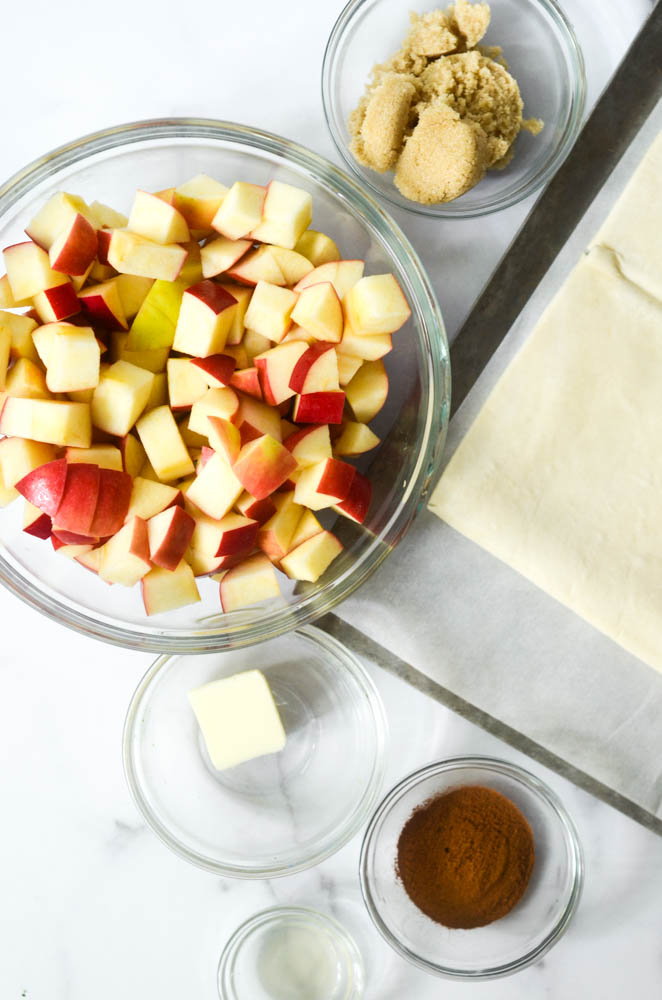 aerial view of ingredients needed: cinnamon, sugar, apples, brown sugar, puff pastry, lemon juice all in bowls.