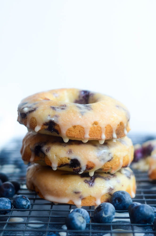 side view of a stack of 3 baked blueberry donuts dipped in glaze with blueberries scattered around