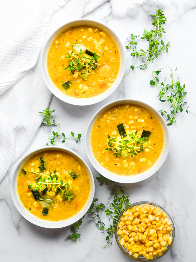 aerial picture of three bowls of corn a and zucchini soup against white marble background with thyme sprigs scattered around