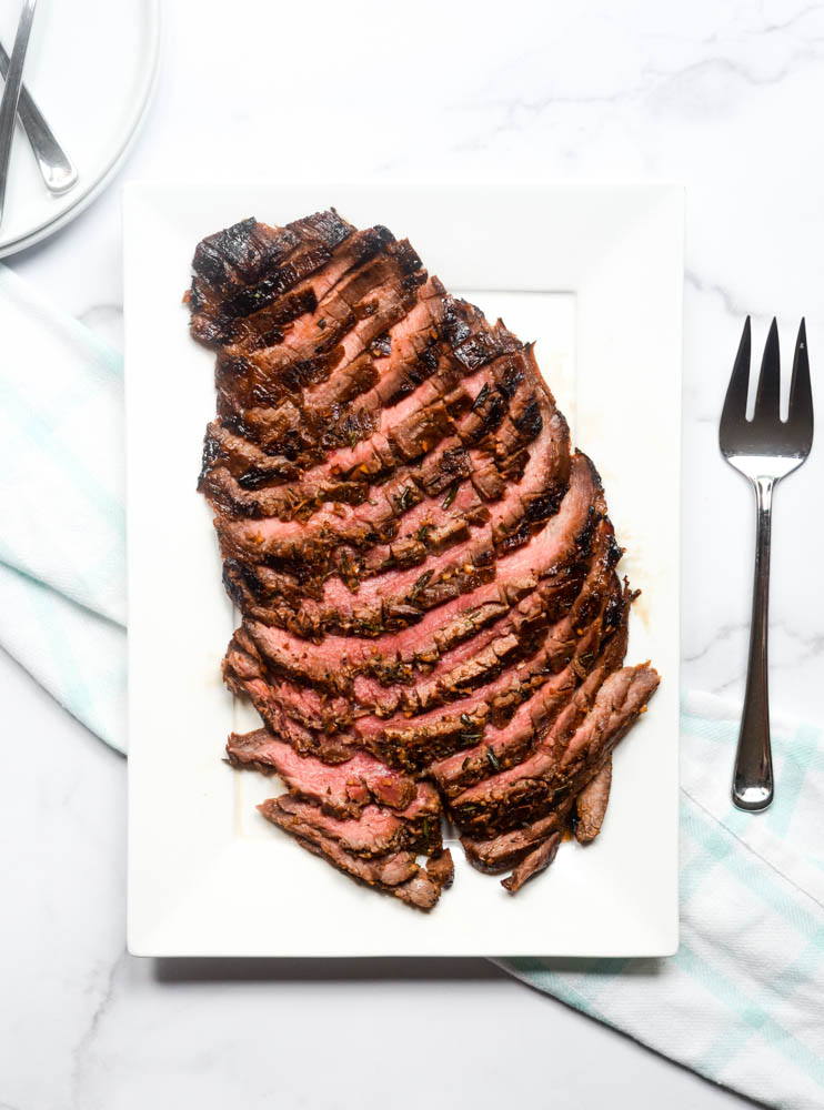 aerial view of white platter with flank steak, plates in corner and serving fork next to platter.