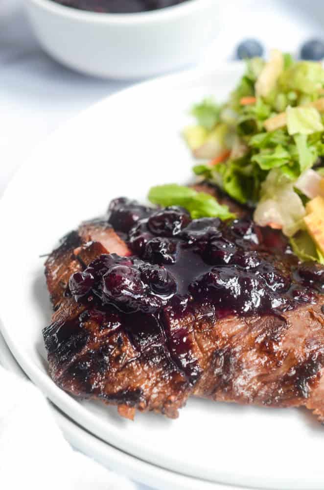 side view of flank steak with blueberry sauce on white plate with salad in background