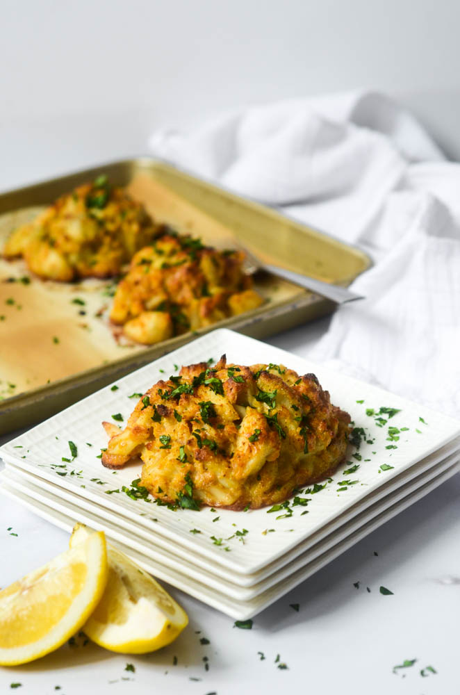 crab cake on stack of white square plates covered with parsley on other crab cakes on baking sheet in background