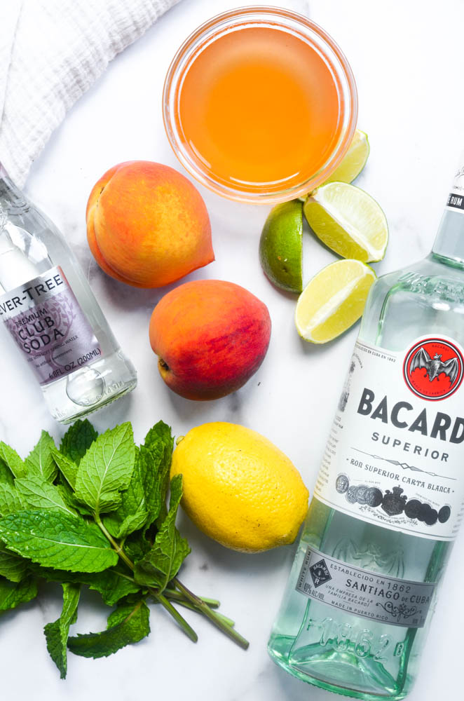 flat lay of ingredients: bowl of peach simple syrup, cut up limes, bacardi, club soda, peaches, lemon, and mint