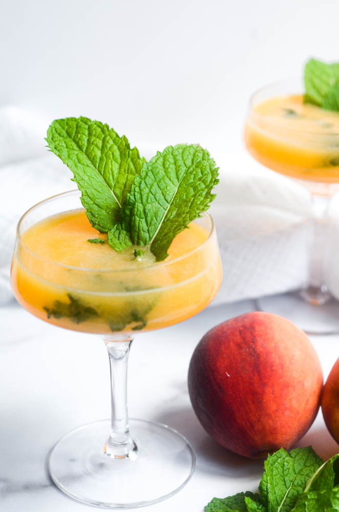 close up of peach mojito with two large mint leaves sticking out.  peaches, limes and mint are scattered around bottom of wine glass.