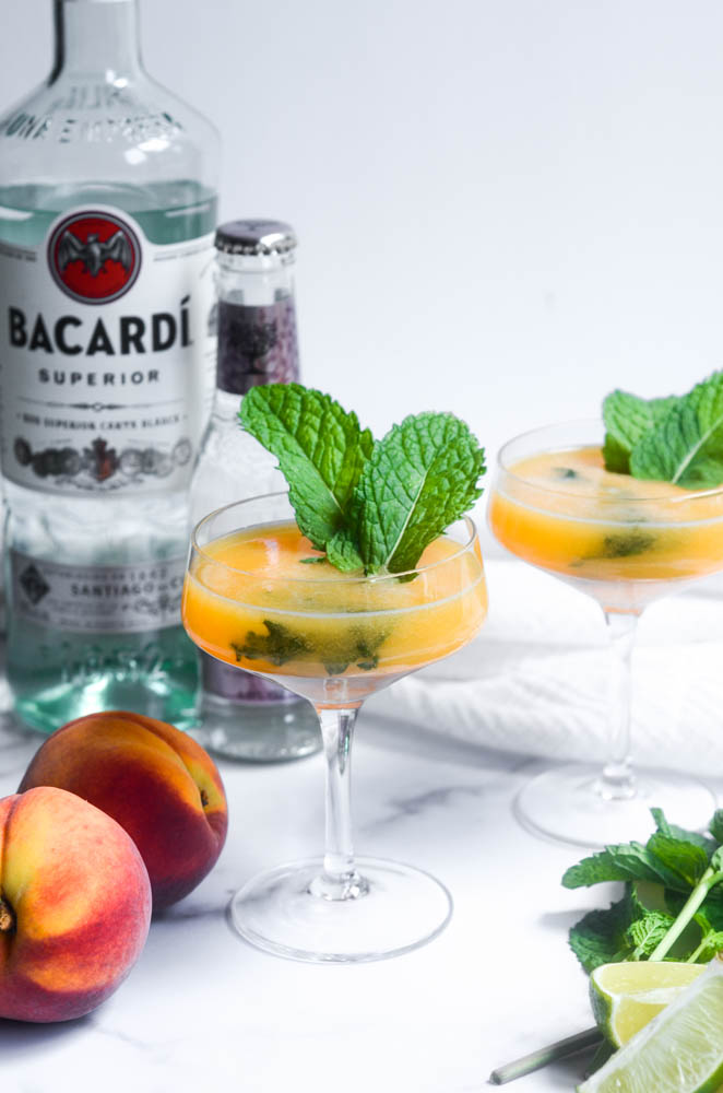 two peach mojitos around peaches, mint, and limes with bottle of bacardi and soda water in the background