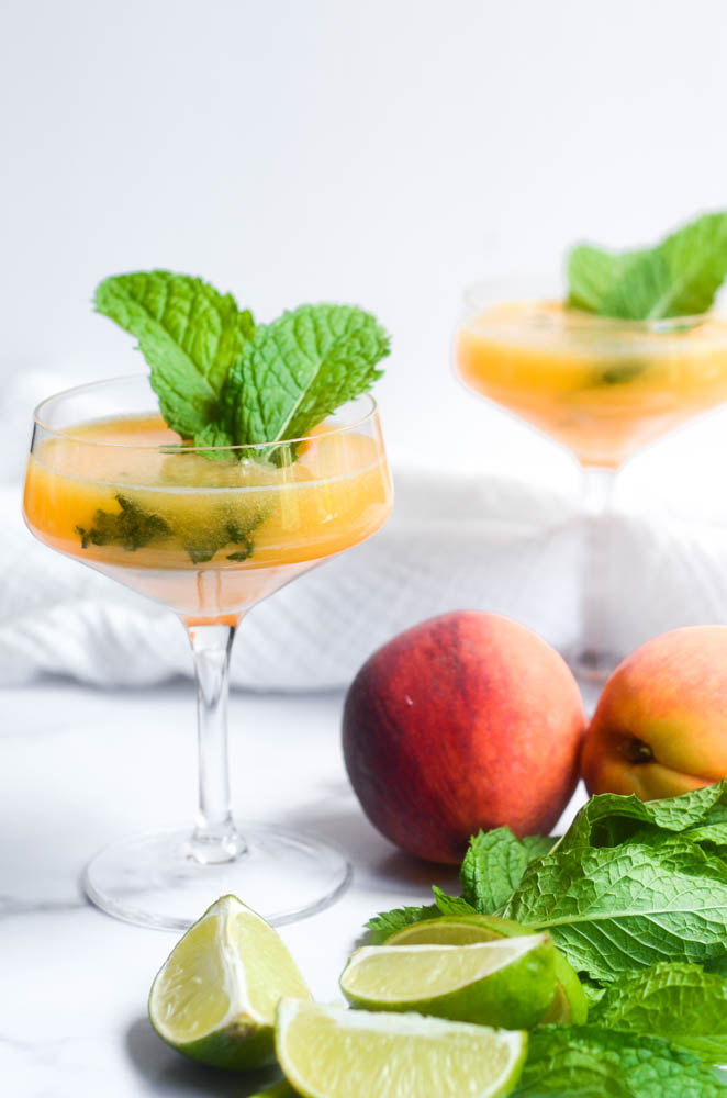 close up of peach mojito in wide mouth wine glass with mint, peaches, and limes in the background