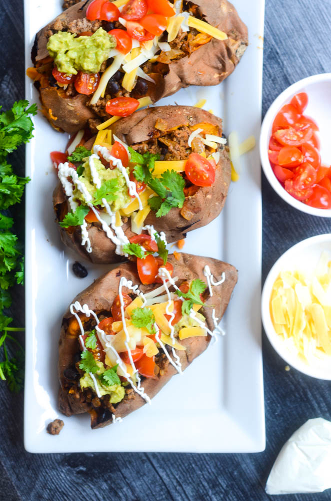 stuffed sweet potatoes on white tray against black backdrop next to bowl of cheese and tomatoes.