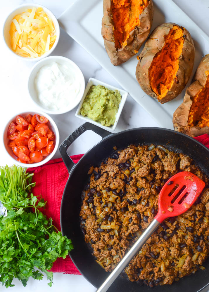 flat-lay of ingredients needed: sweet potatoes, skillet with taco meat in it, and separate bowls of toppings