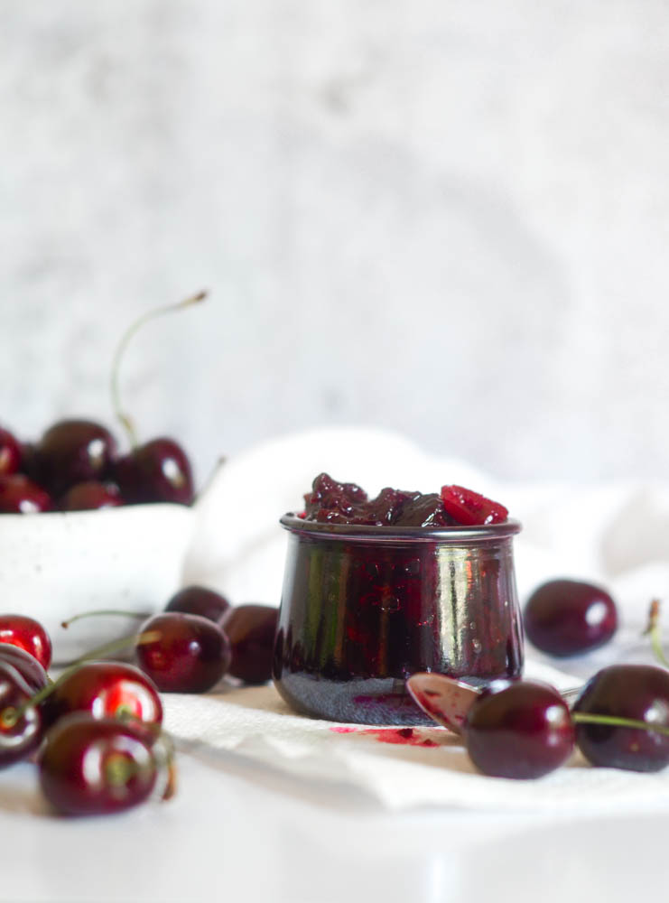 close up of jar of compote surrounded by cherries and bowl of more cherries in background