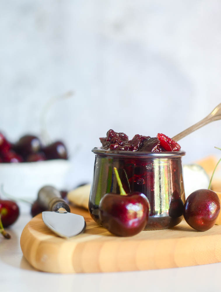 front view of glass jar of compote with cherry on wooden cutting board and cherries and cheese in the background.