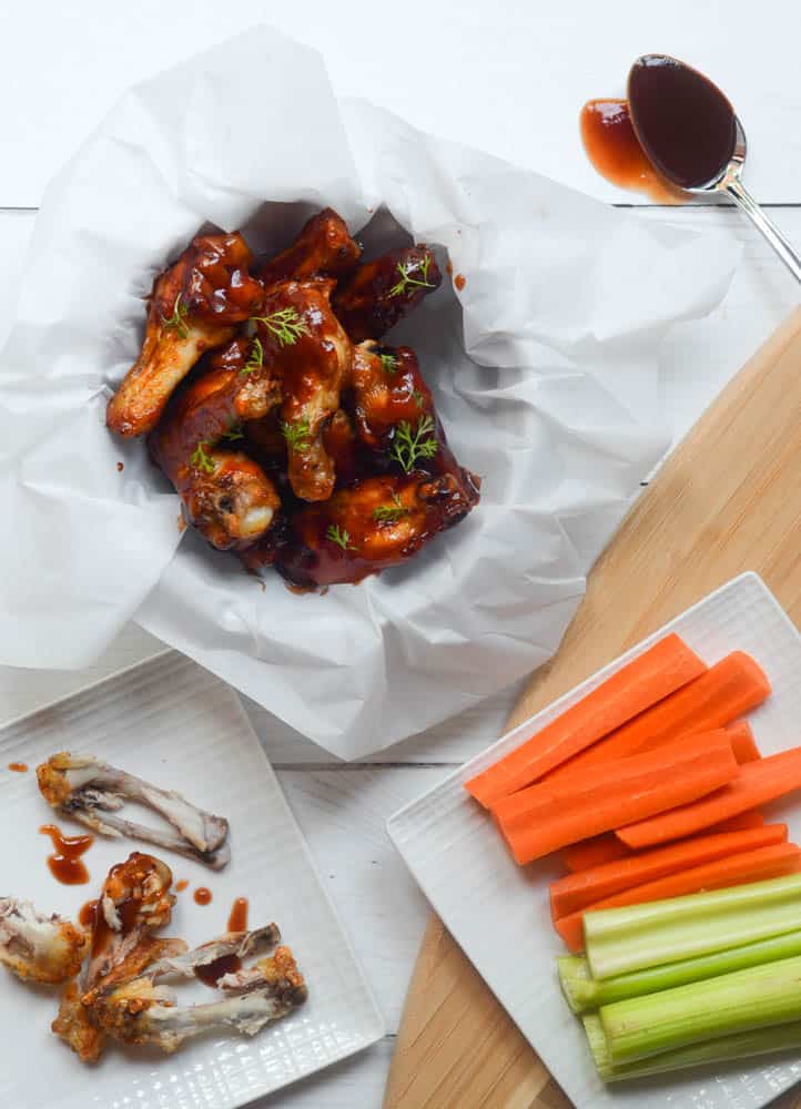 aerial view of bowl o chicken wings in parchment paper nest to square plate with celery and carrots and another plate of chicken wing bones. 