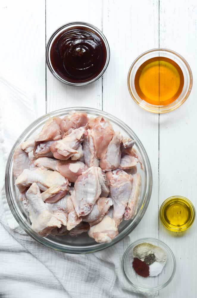 raw chicken wings in bowl with small bowl of spices, olive oil, honey, and BBQ sauce. 