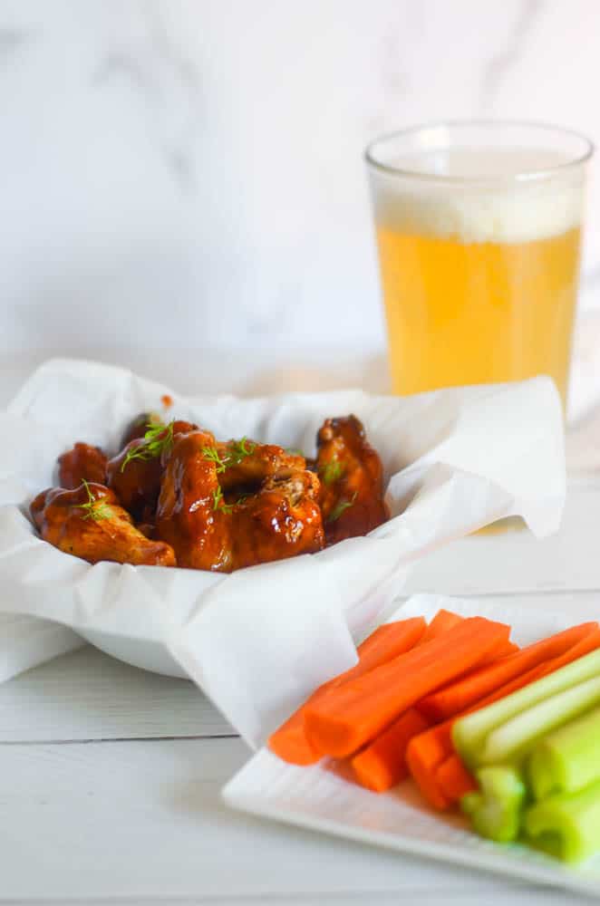 white bowl of chicken wings next to plate of celery and carrots and tall glass of beer. 