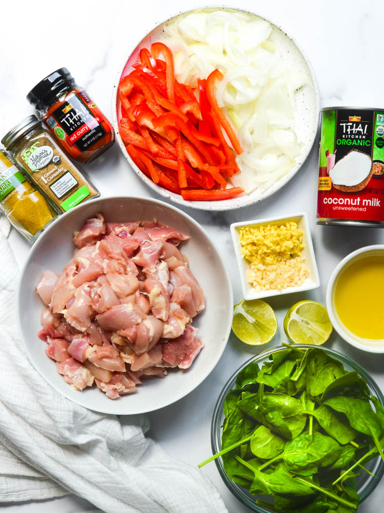 aerial view of ingredients: bowl of onions and red peppers, yellow curry, coriander, red curry paste, coconut milk, bowl of chopped chicken, garlic, ginger, limes, olive oil, and spinach