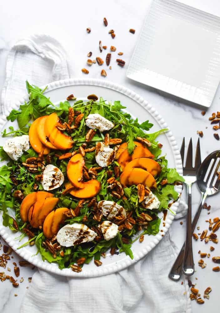 peach burrata salad flatlay on white serving platter next to serving utensils and scattered pecans